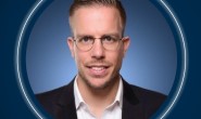 Eine Person mit Brille in einem Anzug vor blauem Hintergrund.