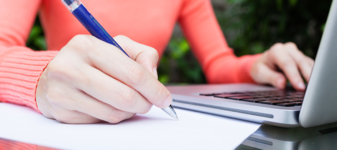 Eine Frau sitzt in einem Garten und schreibt handschriftlich ein Kündigungsschreiben, während sie sich Informationen auf ihrem Laptop ansieht.