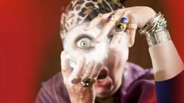 Person mit Schmuck hält eine Lupe vor das Auge, dramatischer Hintergrund.