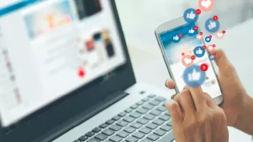 Person scrollt auf Smartphone, Laptop im Hintergrund