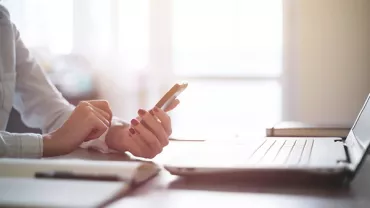 Person hält Smartphone neben Laptop und Notizbuch.