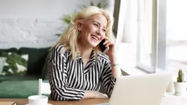 Person mit Telefon am Ohr und Laptop am Tisch, lächelnd.