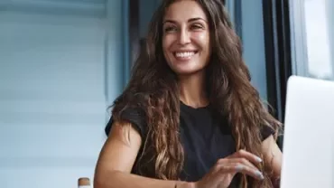 Person am Tisch lächelt und benutzt einen Laptop.