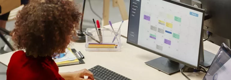 Person arbeitet am Schreibtisch mit Kalender auf dem Computerbildschirm.