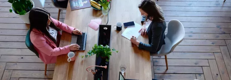 Zwei Personen arbeiten an einem Holztisch, Laptop und Notizen.