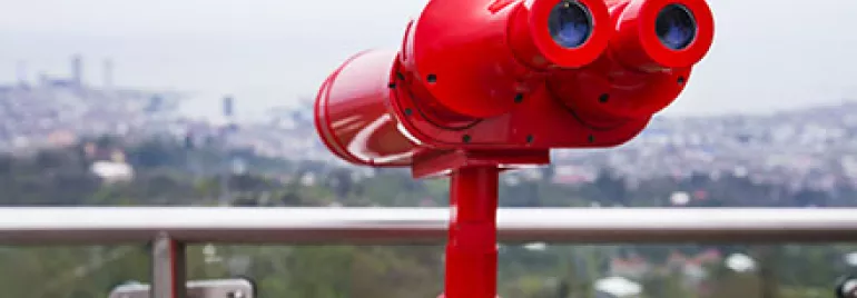 Rotes Fernglas auf einem Aussichtspunkt, mit Stadtblick im Hintergrund.