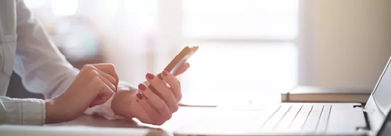 Person hält Smartphone neben Laptop und Notizbuch.