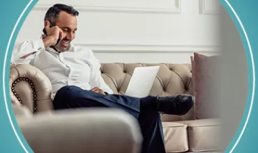Person telefoniert und arbeitet am Laptop auf einem Sofa sitzend.