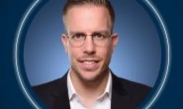 Eine Person mit Brille in einem Anzug vor blauem Hintergrund.