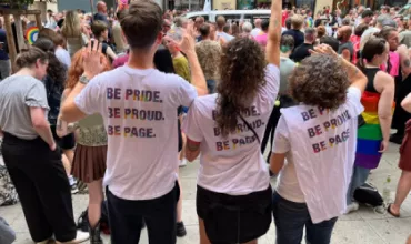 Menschen tragen T-Shirts mit dem Aufdruck 'BE PRIDE. BE PROUD. BE PAGE.' bei einer Parade.